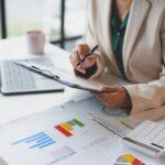 Eine Person in einem Business-Outfit überprüft Finanzunterlagen und Diagramme an einem Schreibtisch, unterstützt von einem Laptop und einem Taschenrechner. Das Bild symbolisiert die professionelle Steuerberatung durch einen Steuerberater in Neu-Ulm zur Optimierung der Finanzen.